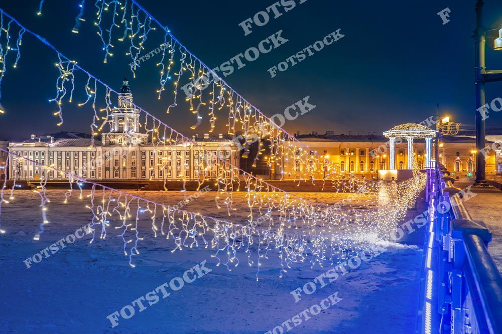 Фотография запечатлела Кунсткамеру, подсвеченную в ночное время, и светящуюся ротонду. На переднем плане гирлянды, украшающие мост, создают сказочный вид. Ледяная поверхность Невы подчеркивает зимний характер снимка. Яркие огни и праздничное освещение дополняют торжественный облик города.